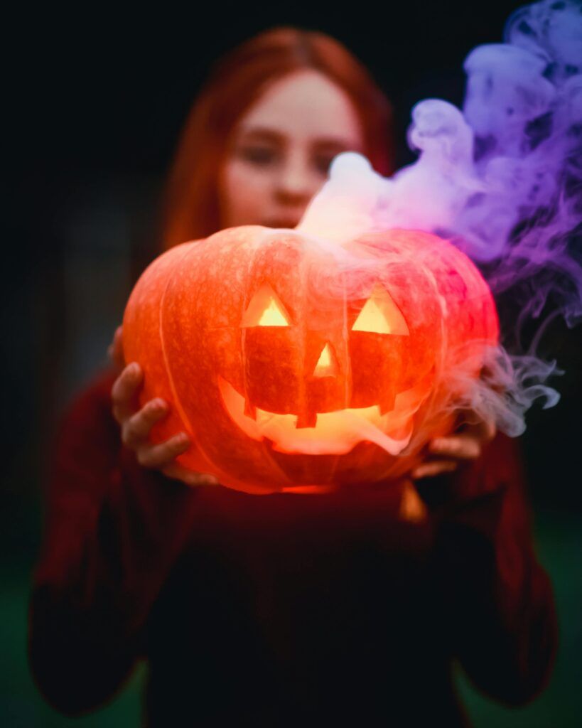 A glowing Jack-o'-lantern with smoke held by an out-of-focus woman, creating a spooky Halloween vibe.